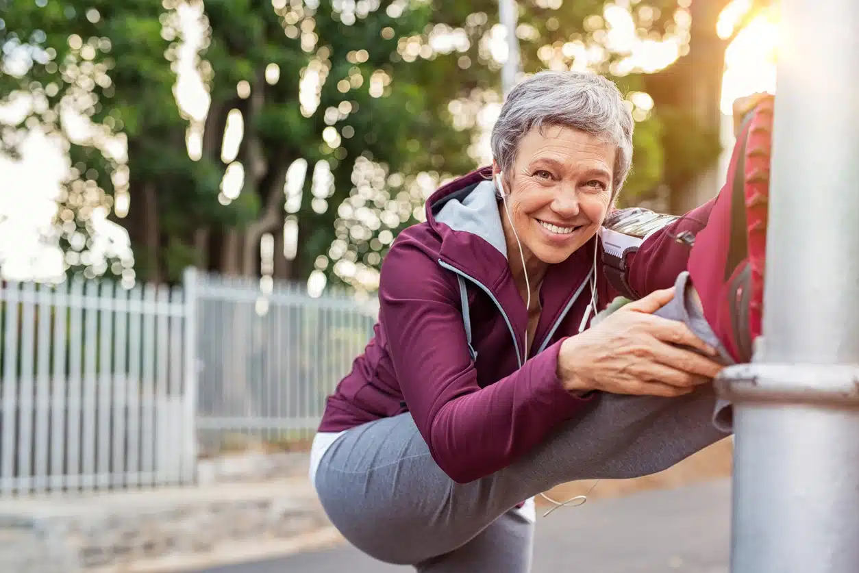 Smiling retired woman listening to music while stretching legs outdoors. Senior woman enjoying daily routine warming up before running at morning. Sporty lady doing leg stretches before workout.